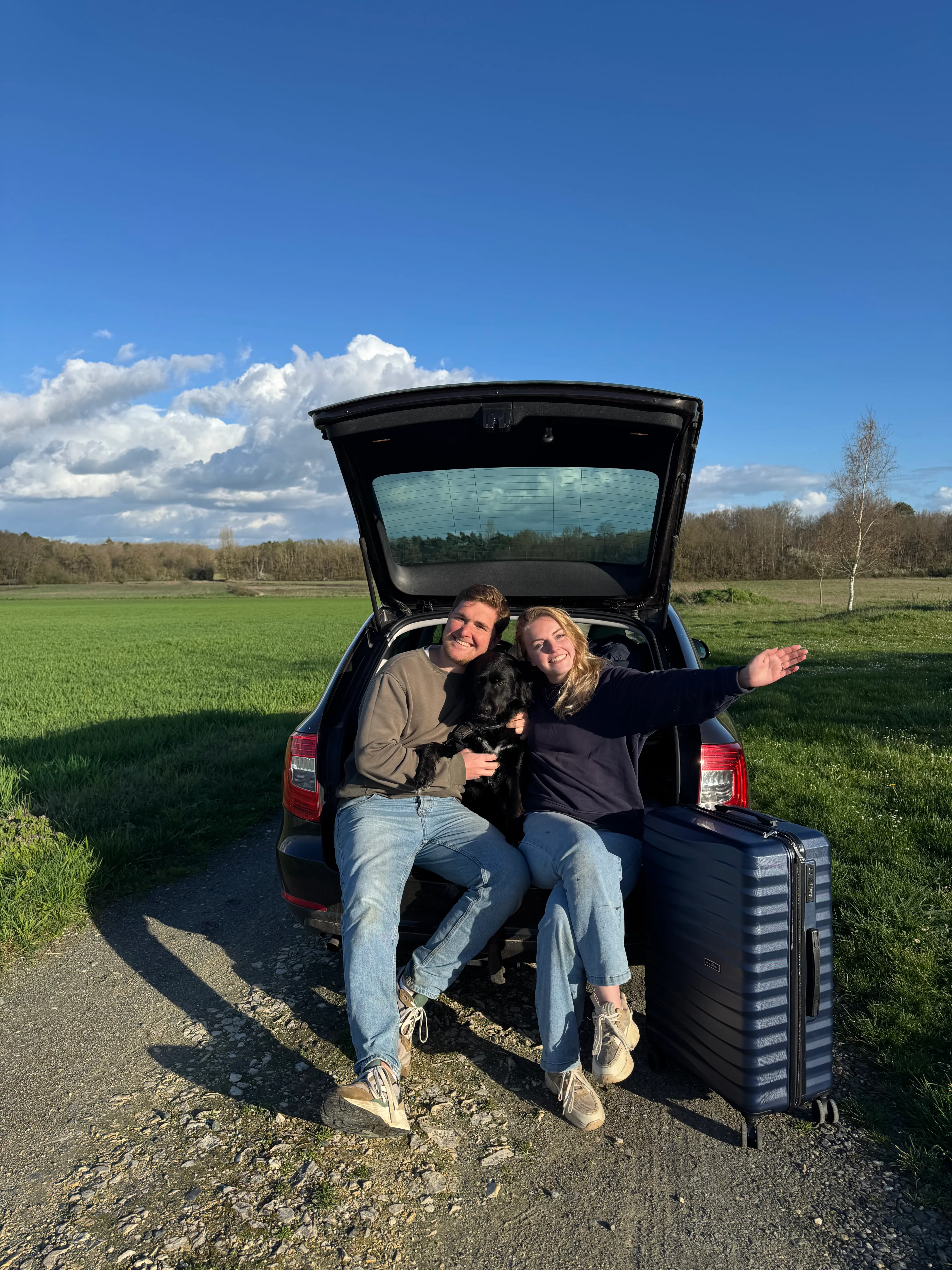Fleur en Laurens zwaaiend in de open kofferbak van hun auto, met hond en koffer, in een Frans landschap